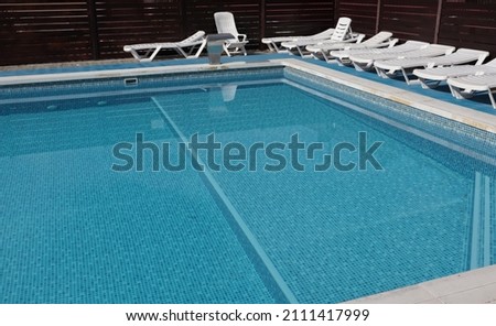 The pool with blue tiles is filled with water and empty white deck chairs.