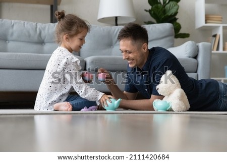 Happy caring millennial dad lying on floor carpet, playing toy tea ceremony with cute small kid daughter, enjoying entertaining activity, spending weekend time together in modern living room. Royalty-Free Stock Photo #2111420684