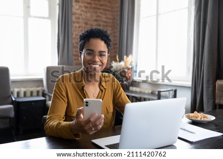 Happy African business woman chatting on digital gadgets online, holding mobile phone, using laptop computer, looking at camera, smiling. Freelance employee at home workplace head shot Royalty-Free Stock Photo #2111420762