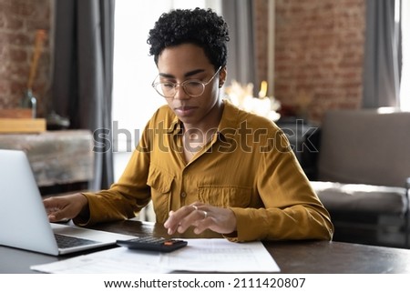 Focused millennial Black business woman calculating finance, money, using calculator, laptop computer at home workplace table, counting budget, paying bills, taxes, rent, mortgage fees Royalty-Free Stock Photo #2111420807