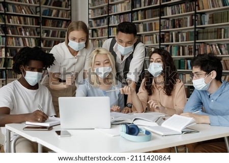 Interracial group of gen Z students wearing facial medical masks sitting at laptop in college library, watching online presentation, discussing learning webinar, working on project together Royalty-Free Stock Photo #2111420864
