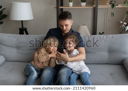 Happy loving young daddy embracing cute small kids siblings, looking together on smartphone screen, enjoying playing games, choosing goods shopping in internet store, modern tech addiction concept.