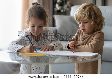 Happy adorable small children siblings drawing pictures in paper album, having fun at glass coffee table at home. Cheerful cute little preschool boy girl friends enjoying creative hobby activity.