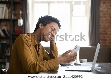 Happy African millennial gadget user girl making video call on smartphone at home office table with laptop. Business woman, student texting message on internet, chatting online on mobile phone Royalty-Free Stock Photo #2111420993