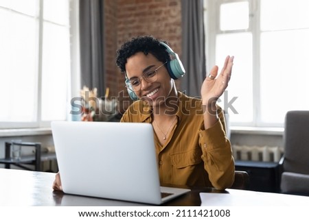 Cheerful happy African American hipster girl in headphones, glasses waving hand hello at webcam of laptop, smiling, speaking on video conference talk, using computer for remote virtual communication Royalty-Free Stock Photo #2111421008