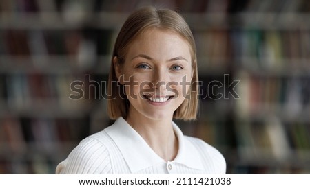 Happy pretty college student girl posing in university library with blurred bookshelves behind. Young millennial woman looking at camera with toothy smile head shot portrait Royalty-Free Stock Photo #2111421038