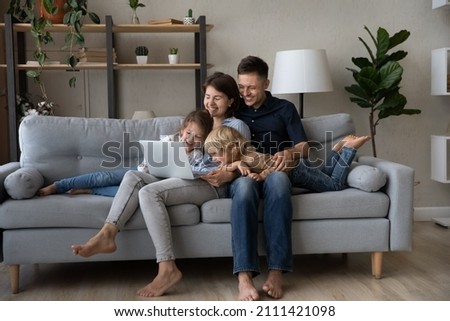Laughing sincere young couple parents and happy small cute kids siblings looking at laptop screen, watching funny movie or comedian movie online, resting on cozy sofa in living room, tech addiction.