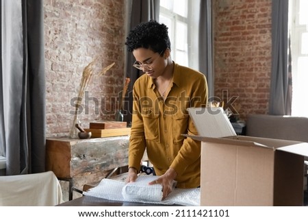 Young African American logistic employee woman packing goods, wrapping, putting books into cardboard carton box, preparing parcel for sending, transportation, collecting donations Royalty-Free Stock Photo #2111421101