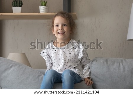 Overjoyed laughing small preschool 6s kid girl having fun, playing on comfortable sofa, enjoying leisure carefree pastime alone in living room, looking in distance watching cartoons on smart TV.