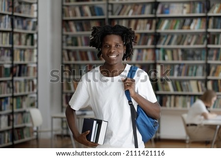 Happy African American student guy with dreads and backpack standing in college library with bookshelves behind, holding stacked books, looking at camera, smiling, Head shot portrait