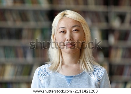 Pensive pretty Asian blonde student girl posing in university library with blurred background behind. Young Chinese hipster woman with pale hair looking at camera head shot portrait