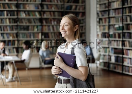 Happy pretty smart college girl walking in public university library, smiling. Thoughtful female student enjoying studying in campus, holding research paper, thinking over learning project