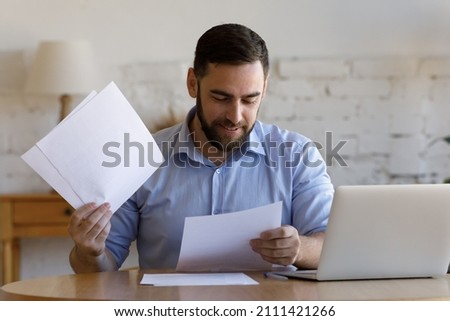 Satisfied smiling business man doing paperwork at home workplace. Entrepreneur reading financial reports, reading documents. Tenant making payment for rent, reviewing bills, bank mortgage notice Royalty-Free Stock Photo #2111421266