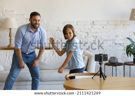 Cheerful excited bloggers dad and joyful active teen son dancing to music in living room, having fun together, posing for smartphone video recording. Active father and kid training, exercising at home