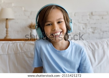 Happy cheerful gen Z school kid boy in big headphones looking at camera with toothy smile, talking on video conference call, studying online from home, attending virtual lesson. Head shot portrait Royalty-Free Stock Photo #2111421272
