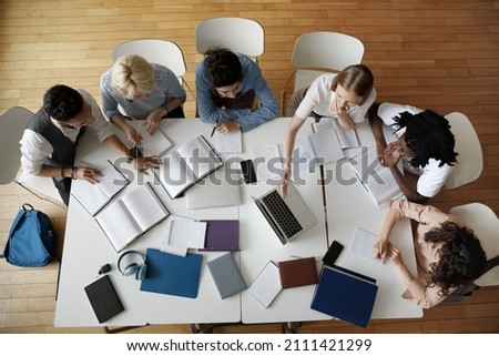 Team of young college students working on study project in university library, using laptop, writing notes at table with books, sharing learning tasks. Top aerial view