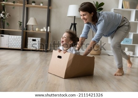 Full length cheerful young mother running barefoot on heated floor, pushing box with seated little adorable laughing kid daughter, having fun together at home, daycare, family relations concept. Royalty-Free Stock Photo #2111421326