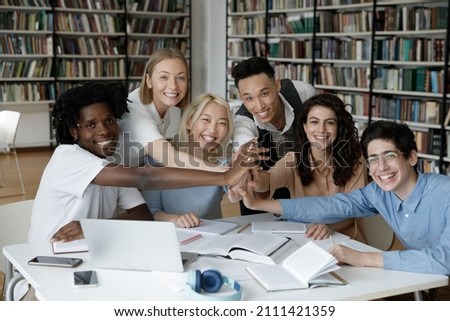 Mixed race team of happy diverse college students giving group high five over table with books in library, looking at camera, smiling. Multiethnic group of teens enjoying learning teamwork portrait