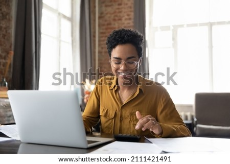Happy young Afro American entrepreneur woman in glasses counting profit, on calculator at laptop computer, analyzing benefits, enjoying financial success, job high result, smiling Royalty-Free Stock Photo #2111421491