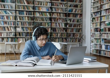 Focused nerdy smart student in glasses and headphones working hard on research study project in library, watching online learning video tutorials, conference, writing notes at pile of books