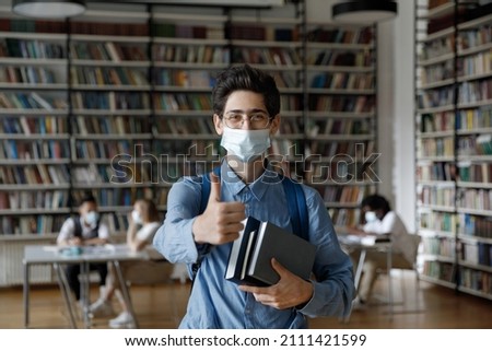 Happy fresh college student covering breathing organs with facial mask, holding books, showing like thumb up gesture in library, recommending protection from viral infection, covid