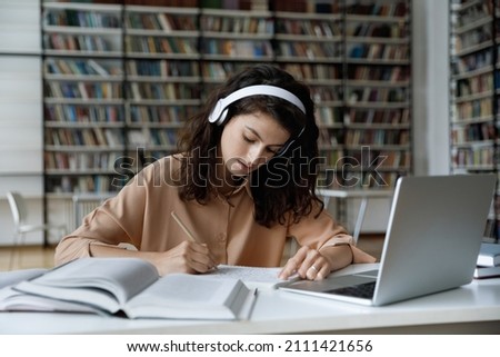 Serious busy hardworking student girl in headphones working essay, study project in college public library, attending online learning conference, watching video lesson on internet, writing notes