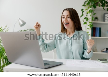 Young excited fun successful employee business woman 20s in blue shirt do winner gesture sit work at workplace white desk with laptop pc computer at modern office indoors. Achievement career concept.