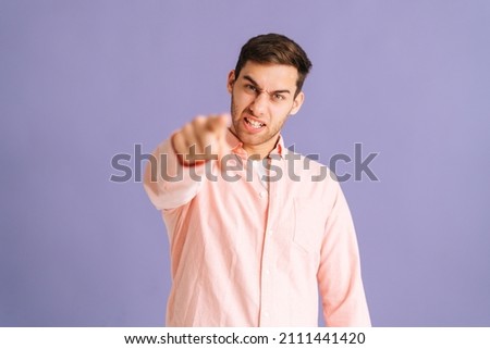 Portrait of angry handsome young man pointing index finger at you on purple isolated background, looking at camera. Annoyed strict guy pointing index finger on camera choosing you. Royalty-Free Stock Photo #2111441420