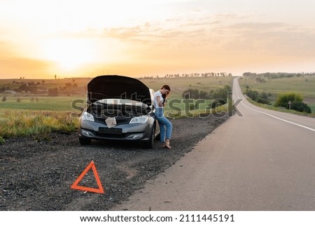 A frustrated young girl stands near a broken-down car in the middle of the highway during sunset. Breakdown and repair of the car. Waiting for help. Car service. Car breakdown on road. Royalty-Free Stock Photo #2111445191