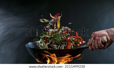 Freeze Motion of Wok Pan with Flying Ingredients in the Air and Fire Flames. Royalty-Free Stock Photo #2111499230