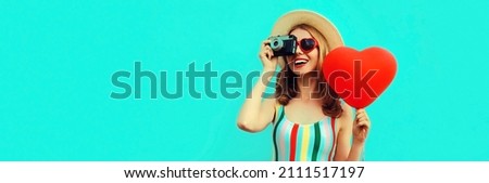 Summer portrait of happy smiling young woman with retro camera and red heart shaped balloon wearing a straw hat on blue background