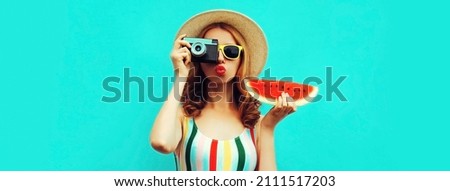 Summer portrait of stylish young woman with retro film camera and slice of fresh watermelon wearing straw hat posing on blue background