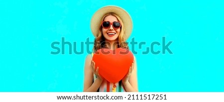 Portrait of happy smiling young woman with big red heart shaped balloon having fun wearing a summer straw hat, sunglasses on blue background