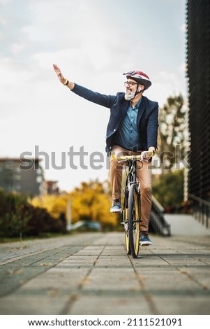 Successful middle-aged businessman waving to someone while riding bicycle on his way to work through the city. Royalty-Free Stock Photo #2111521091