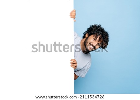Positive Indian or Arabian guy, in casual t-shirt, peeking out from behind advertisement whiteboard, demonstrating blank copy space for your text or design, isolated blue background, mock-up concept Royalty-Free Stock Photo #2111534726