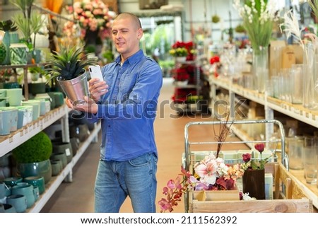 Man buyer holding pot with replica plant and taking pictures with his smartphone while shopping in home goods store.