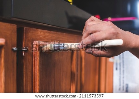 A handyman paints a fresh coat of varnish on the surface of a base kitchen cabinet with a medium sized brush. Home renovation or finishing works. Royalty-Free Stock Photo #2111573597