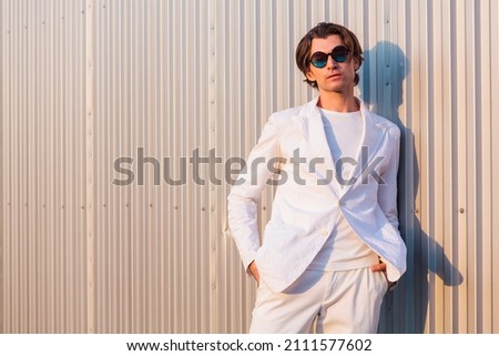 Portrait of a handsome man dressed in a white suit and round hippie sunglasses standing near the wall during sunset