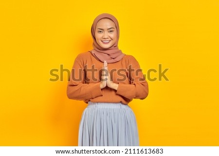 Portrait of smiling young Asian Muslim woman in brown sweater looking confident, open sign to greet and greet customers isolated over yellow background