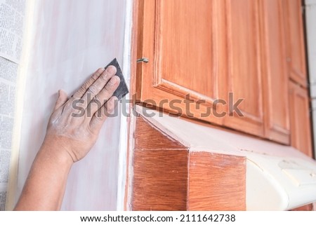 A handyman using a piece of sandpaper to smoothen out the edge of a wall cabinet prior to painting. Home renovation or finishing works. Royalty-Free Stock Photo #2111642738
