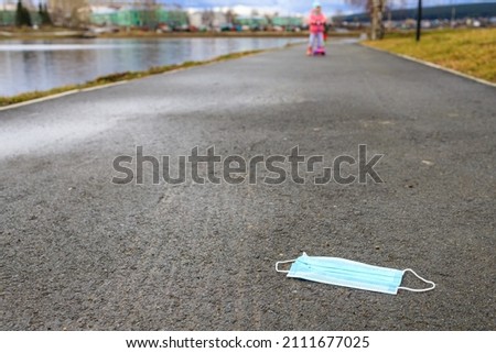 a medical mask in the foreground is lying on the sidewalk. background child on a scooter on the sidewalk. The baby is blurry out of focus