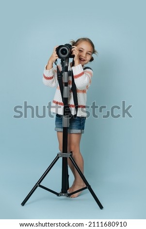 Cheerful little girl in striped sweater standing behind a digital camera mounted high on a tripod over blue background. She's looking at the camera.