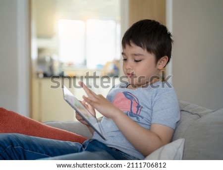 Kid sitting on sofa watching cartoon or playing game on tablet, Child boy using digital pad learning lesson online on internet, Home schooling,Distance learning online education concept