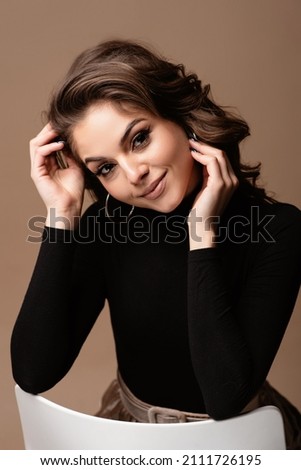 Beautiful seductive young european woman portrait. Girl wear black sweater with beautiful face and hair sitting on chair looking at camera. Isolated on brown background Royalty-Free Stock Photo #2111726195