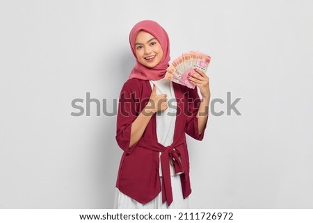 Cheerful beautiful Asian woman in casual shirt and hijab holding Indonesian rupiah banknotes, showing thumb up gesture isolated over white background. People religious lifestyle concept Royalty-Free Stock Photo #2111726972