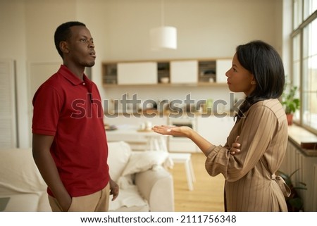 Quarrel. Young ethnicity couple having unpleasant conversation against kitchen background, man in red polo t-shirt with offended look facing his girlfriend, girl asking him where he's been last night Royalty-Free Stock Photo #2111756348