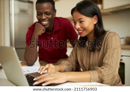 Close-up picture of two enthusiastic dark-skinned people in kitchen, young attractive, cheerful, natty woman sitting in front of laptop, typing, African man in red, watching her working smiling widely