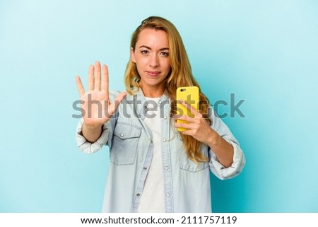 Caucasian woman holding mobile phone isolated on blue background standing with outstretched hand showing stop sign, preventing you.