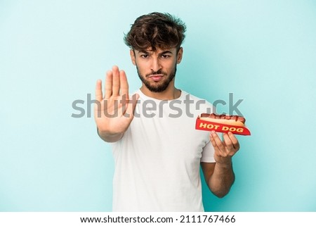 Young arab man holding a hotdog isolated on blue background standing with outstretched hand showing stop sign, preventing you.