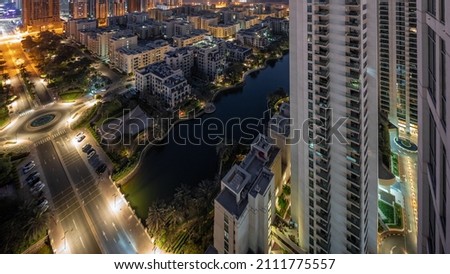 Pond and low rise buildings in Greens district aerial night to day transition timelapse. Dubai skyline with skyscrapers in Barsha Heights district on a background